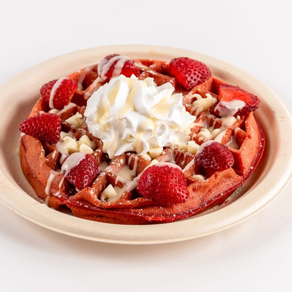 Red velvet waffle topped with whipped cream, strawberries, white chocolate chips, and syrup.