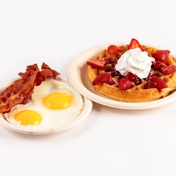 A Waffles Café breakfast with a waffle topped with strawberries and whipped cream, and a side of bacon and fried eggs.