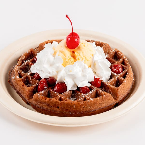 Chocolate waffle topped with vanilla ice cream, whipped cream, cherries, and powdered sugar.